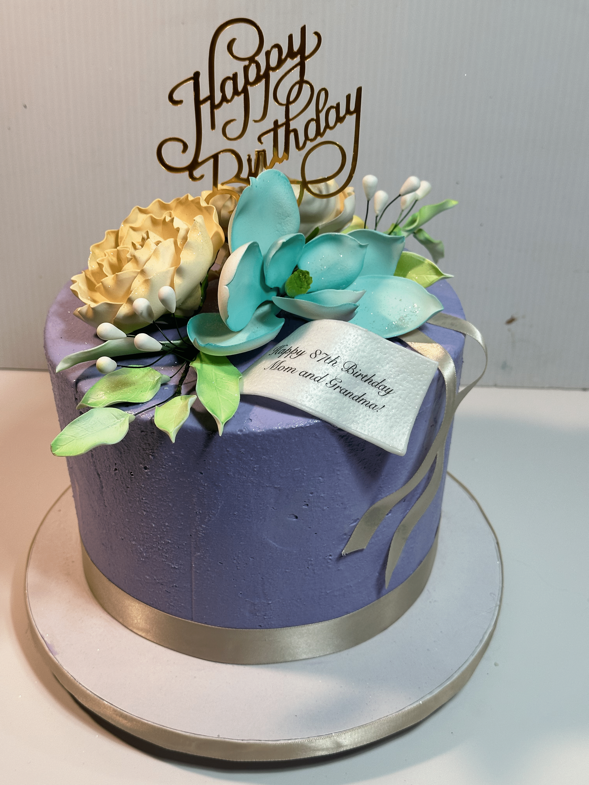 Lavender 87th birthday cake topped with teal and cream sugar flowers and a gold topper
