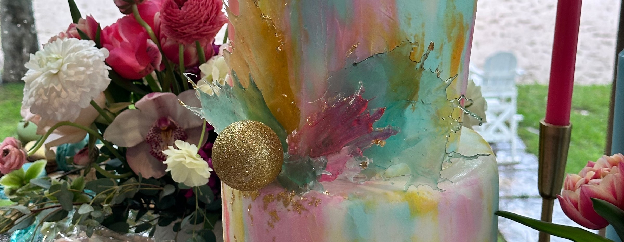 Watercolor-painted tiered cake with a gold sphere beside pink and white fresh flowers