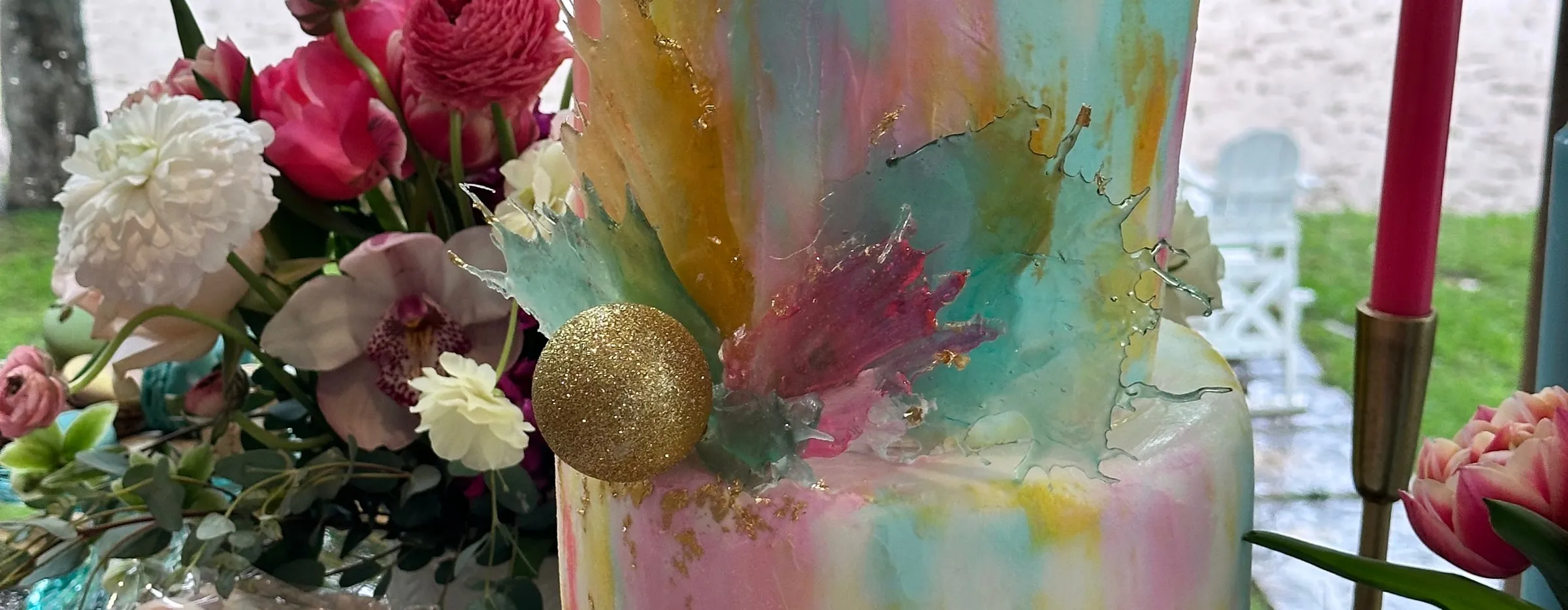 Watercolor-painted tiered cake with a gold sphere beside pink and white fresh flowers